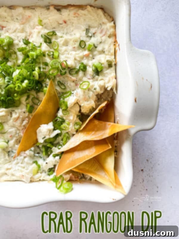Savory Crab Rangoon Dip 14 A beautifully presented warm Crab Rangoon Dip in a baking dish, garnished with green onions and surrounded by wonton chips on a rustic wooden board.