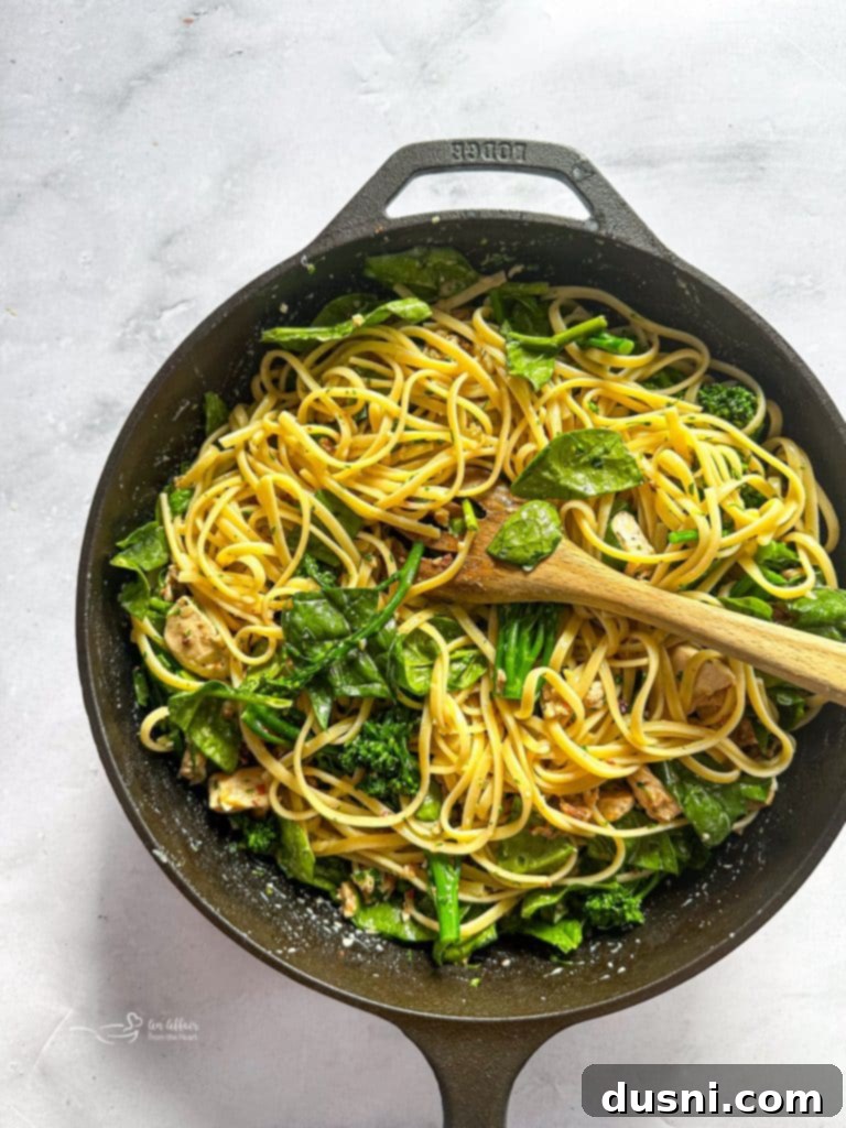 A large skillet with creamy carbonara sauce, chicken, and bacon, with cooked pasta, broccolini, and spinach about to be added.