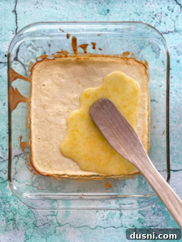 thick lemon glaze being poured and spread evenly over the baked and cooled lemon brownies in the pan