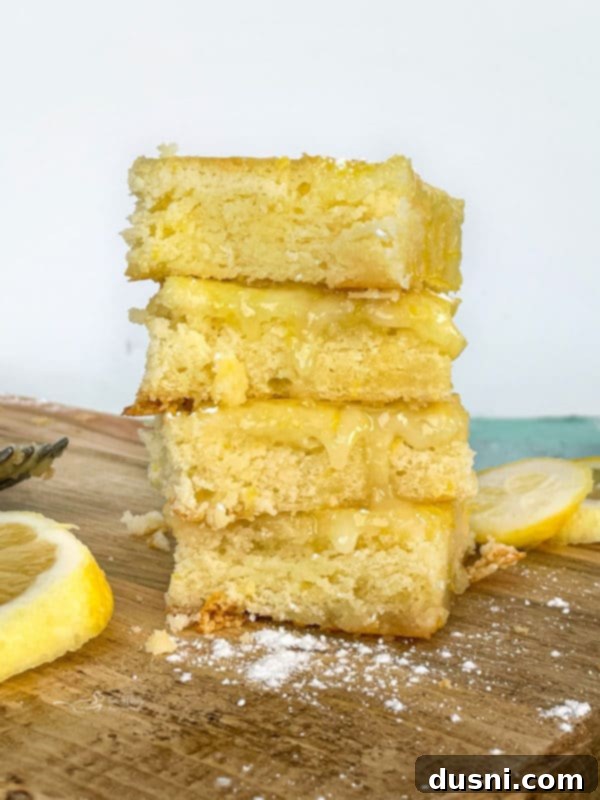 front stack of three lemon brownies on a white surface with lemons in the background, showcasing their bright color