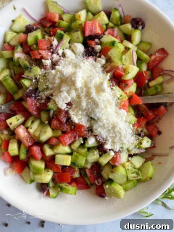 Greek cucumber salad in a white bowl, now topped with generous crumbles of feta cheese, ready for final seasoning.
