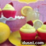 Zesty Lemon Cupcakes with Raspberry Burst 2 Side view Lemonade cupcakes on a yellow plate and on a white cake platter with half of lemons and a glass of lemonade on a pink surface. text "lemonade cupcakes with raspberry filling"