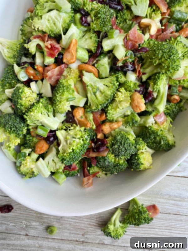 Smoky Cashew Broccoli Crunch 2 Top view of white bowl filled with bacon cashew broccoli salad, showcasing fresh broccoli florets, cashews, raisins, and crumbled bacon, all coated in a creamy dressing.