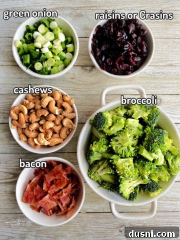Smoky Cashew Broccoli Crunch 4 Wooden surface displaying fresh ingredients for broccoli salad: cashews, green onions, raisins, fresh broccoli florets, and strips of raw bacon.