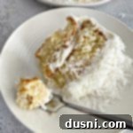 A fork with a bite of the COCONUT CREAM POKE CAKE WITH COCONUT WHIPPED CREAM FROSTING next to the slice of cake on a white plate