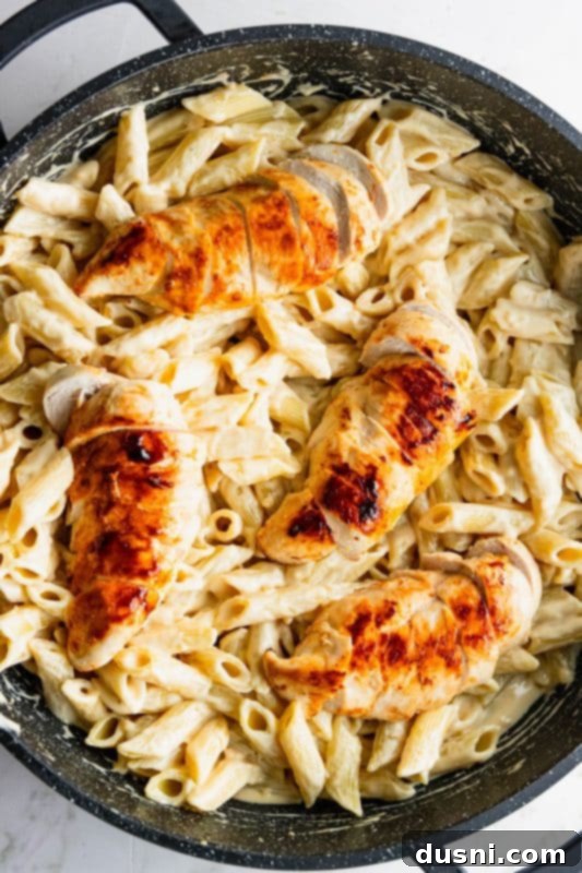 Sliced cooked chicken breasts being placed on top of the creamy penne pasta in the skillet.