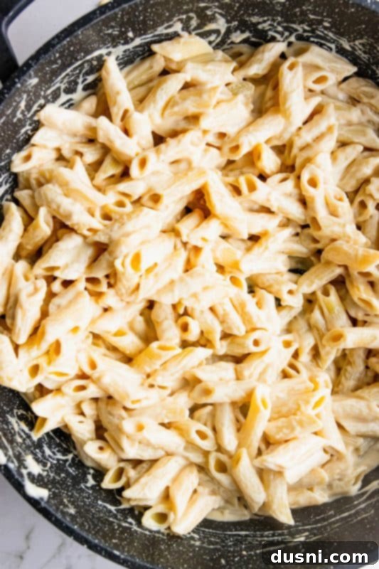 Cooked penne pasta being added to the creamy sauce in a large skillet.