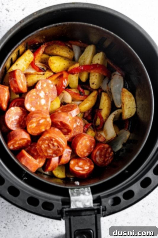 Adding sliced sausage to the air fryer basket with potatoes and vegetables