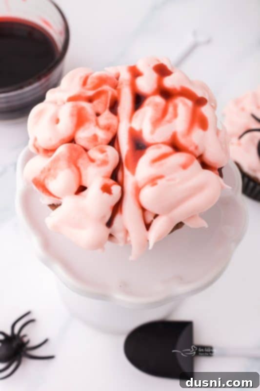 Ghoulish Grey Matter Cupcakes 2 A beautifully decorated Halloween Brain Cupcake, showcasing the pink cream cheese frosting shaped like a brain, with realistic edible blood drizzled over it. The cupcake sits on a plain background, highlighting its ghoulish details, perfect for a spooky Halloween treat.