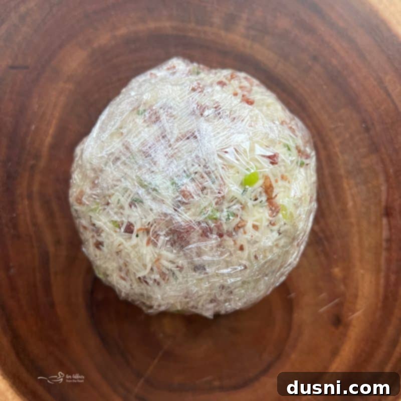 The cheese ball, partially coated with the herb, bacon, and cheese mixture, being rolled on plastic wrap to fully coat.