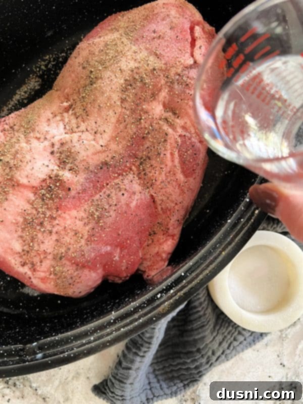 Water being poured into the bottom of a roasting pan with a seasoned pork roast.