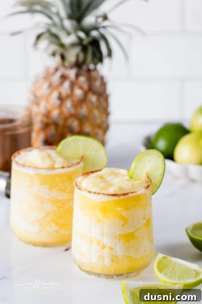 Two glasses of Frozen Pineapple Margaritas, one with a lime wedge garnish, on a rustic wooden surface.