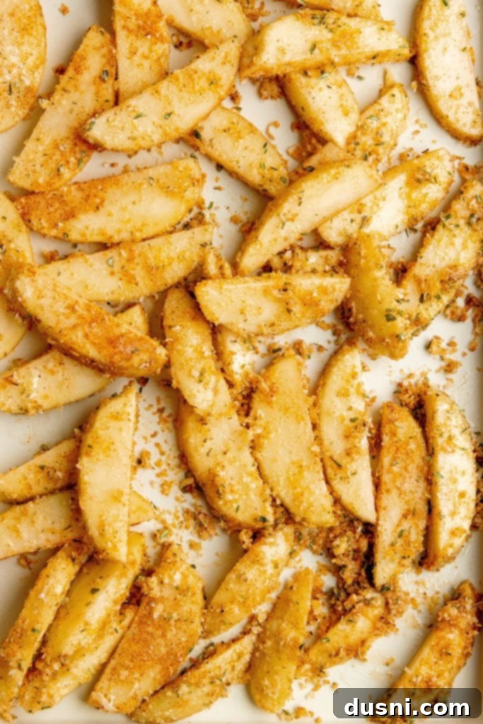 Potato wedges, seasoned and coated with parmesan, spread evenly on a baking sheet.