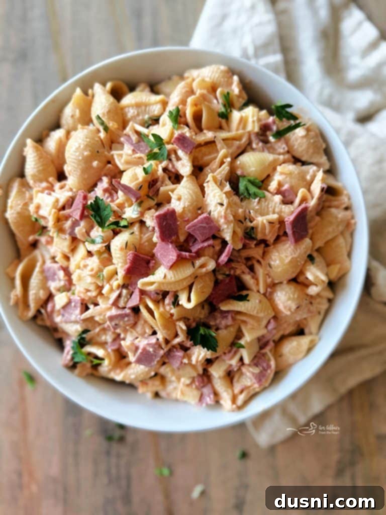 Reuben Pasta Salad in a large white bowl with a wooden serving spoon, garnished with caraway seeds and parsley.